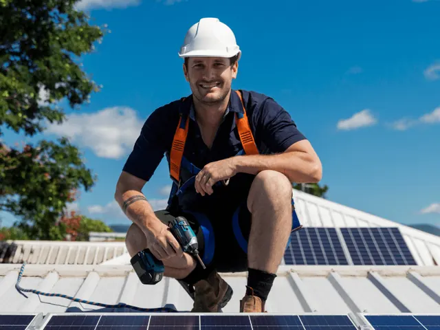 Smiling installer in a white hard hat crouching beside solar panels on a roof