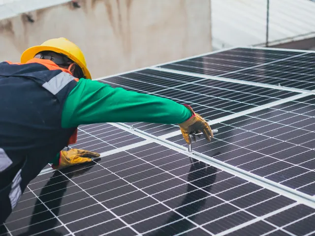 Technician in a yellow hard hat and green sleeve reaching across solar panels